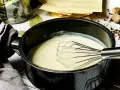 Cremige béchamelsauce mit Sahne wird in einem Topf mit einem Schneebesen zubereitet. Daneben liegen Lasagneplatten und Gewürze.