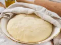 Der weltbeste Pizzateig mit Trockenhefe ruht abgedeckt in einer Schüssel. Daneben liegen Salz, Olivenöl und ein Nudelholz.