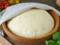 Ein aufgegangener Pizzateig in einer Holzschüssel, bedeckt mit einem Tuch. Die Frage "wie lange pizzateig gehen lassen" wird hier visuell beantwortet.