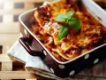 Herzhafte Lasagne Bolognese, eine der beliebtesten Lasagne Arten, dampft in einer braunen Auflaufform mit weißen Punkten. Frisch garniert mit Basilikum.