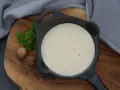 Cremige Bechamelsauce in einer Gusseisenpfanne, bereit für die Lasagne. Muskatnuss und Petersilie deuten auf die feinen Aromen hin.