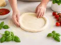 Hände formen einen Pizzaboden nach einem Pizzateig Rezept. Frische Tomaten und Basilikum liegen bereit.