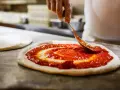 Eine Hand verteilt Tomatensauce auf einem Teigboden, die Vorbereitung für eine authentische pizza napoli original.