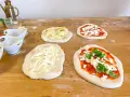 Vier vorbereitete Pinsa-Teiglinge liegen auf einem Holztisch. Einer mit Apfelscheiben, einer mit Tomatensoße und Mozzarella, einer nur mit Mozzarella und einer mit Tomatensoße, Mozzarella und Basilikum.