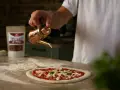 Ein Mann übergießt mit Olivenöl einen **Waldi Pizzateig**. Im Hintergrund eine Tüte mit Chiliflocken.
