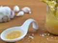 Ein Löffel mit einer köstlichen Knoblauchpaste, die an italienische Aromen erinnert. Daneben eine Flasche mit einer gelben Flüssigkeit und Knoblauchzehen.