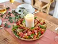 Ein festliches Arrangement mit italienischen Snacks zum Wein: Salami, Prosciutto, Käse, Oliven und Brot, umrahmt von Kräutern und einer Kerze.