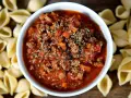 Herzhafte pancetta bolognese in einer Schüssel, umgeben von ungekochten Muschelnudeln.