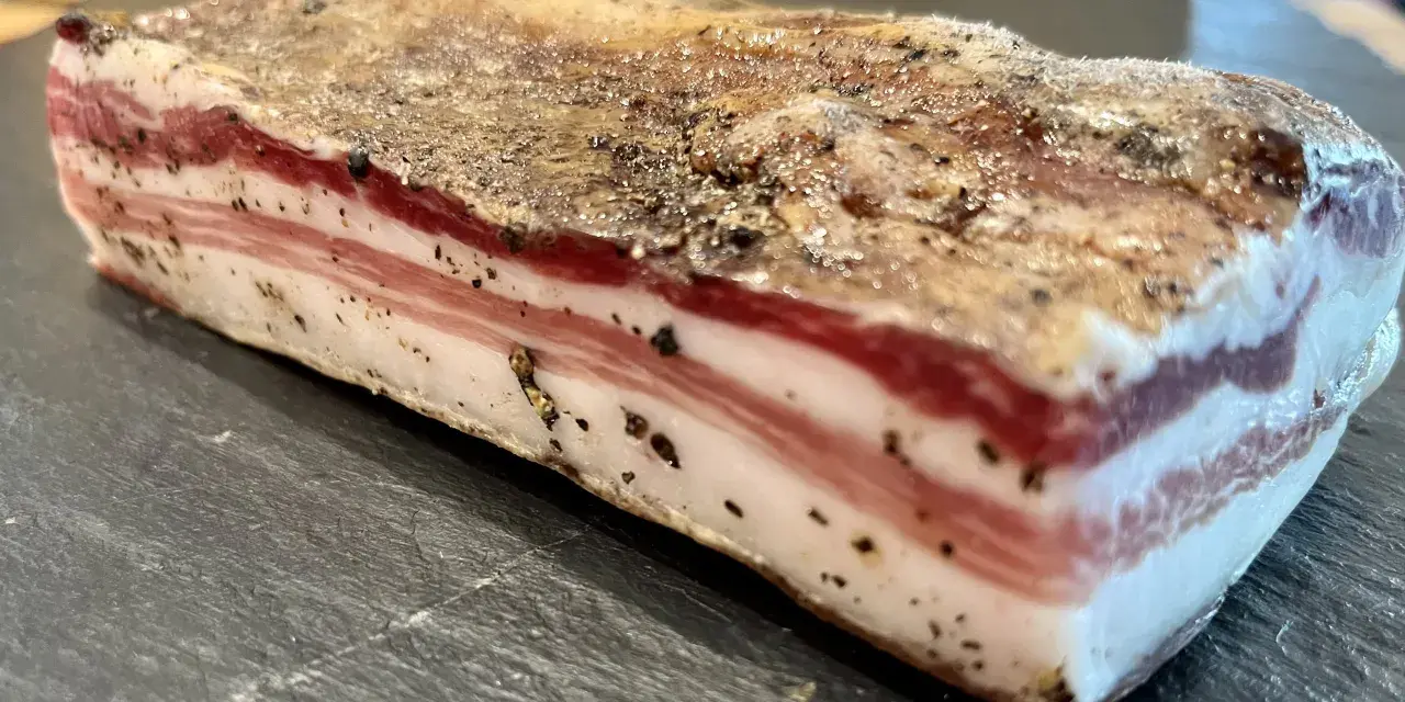 Ein Stück roher Pancetta mit deutlichen Schichten aus Fleisch und Fett, bestreut mit schwarzem Pfeffer. Perfekt, um ihn roh zu genießen.