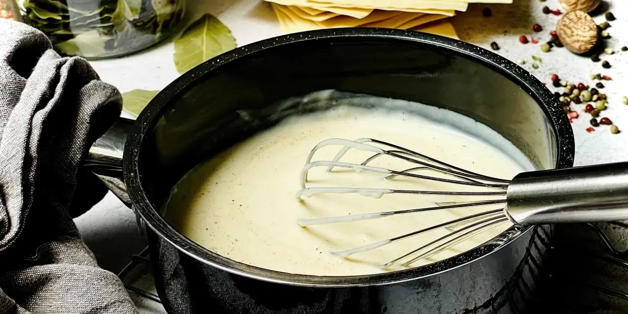 Cremige béchamelsauce mit Sahne wird in einem Topf mit einem Schneebesen zubereitet. Daneben liegen Lasagneplatten und Gewürze.