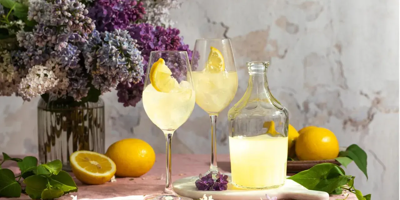 Erfrischender Limoncello Spritz mit Bitter Lemon, garniert mit Zitronenspalten, neben frischen Zitronen und einem üppigen Fliederstrauß.