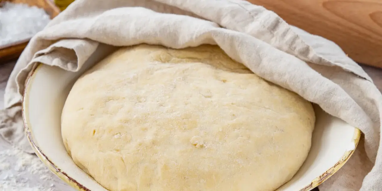 Der weltbeste Pizzateig mit Trockenhefe ruht abgedeckt in einer Schüssel. Daneben liegen Salz, Olivenöl und ein Nudelholz.