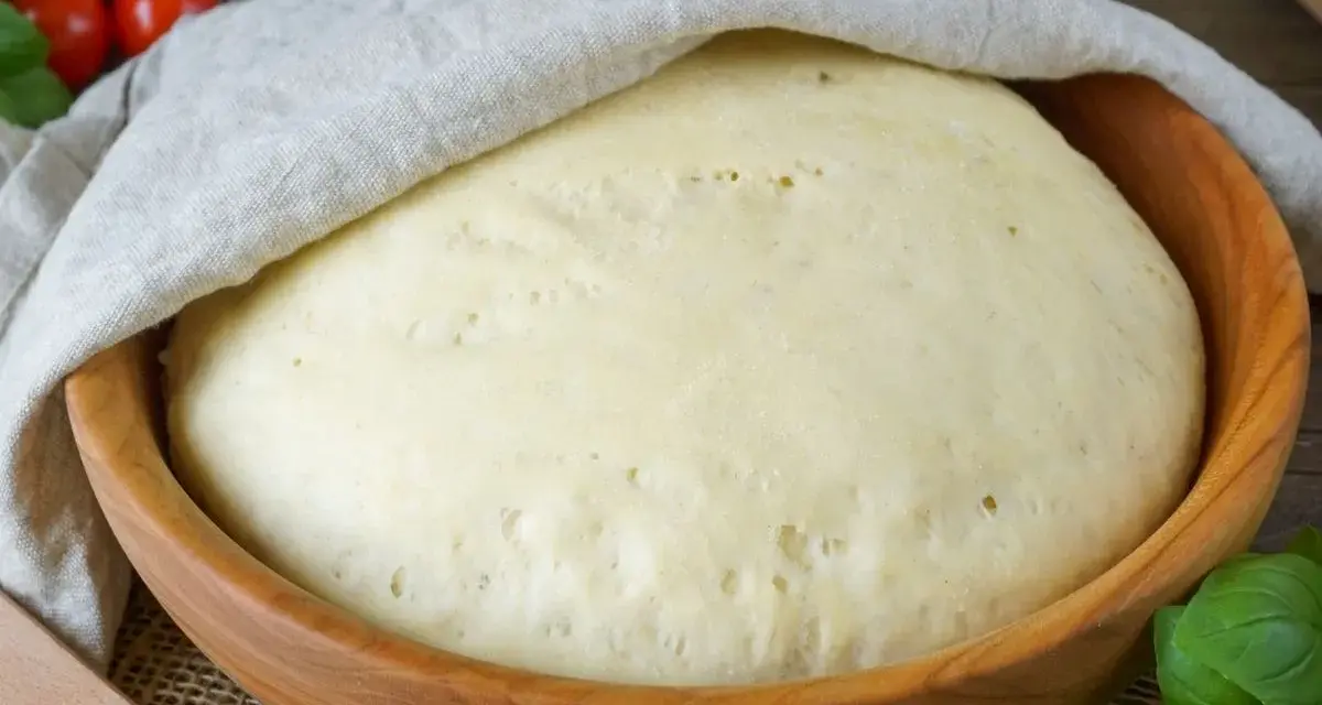Ein aufgegangener Pizzateig in einer Holzschüssel, bedeckt mit einem Tuch. Die Frage "wie lange pizzateig gehen lassen" wird hier visuell beantwortet.