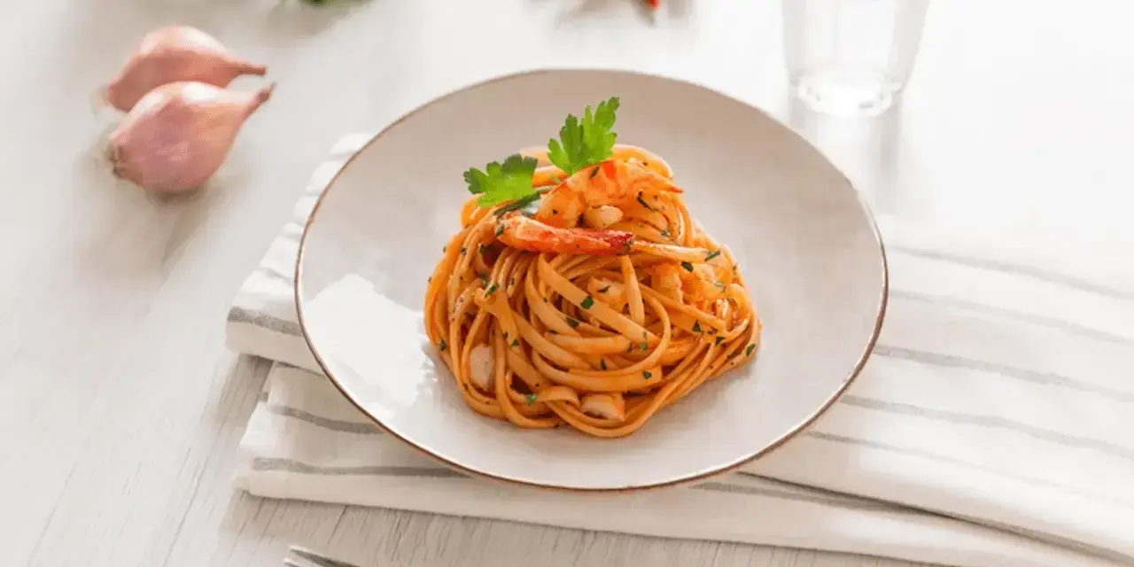 Ein Teller mit Linguini Nudeln mit Garnelen und Tomatensauce, garniert mit Petersilie. Daneben Schalotten, Chilis und ein Glas Wasser.