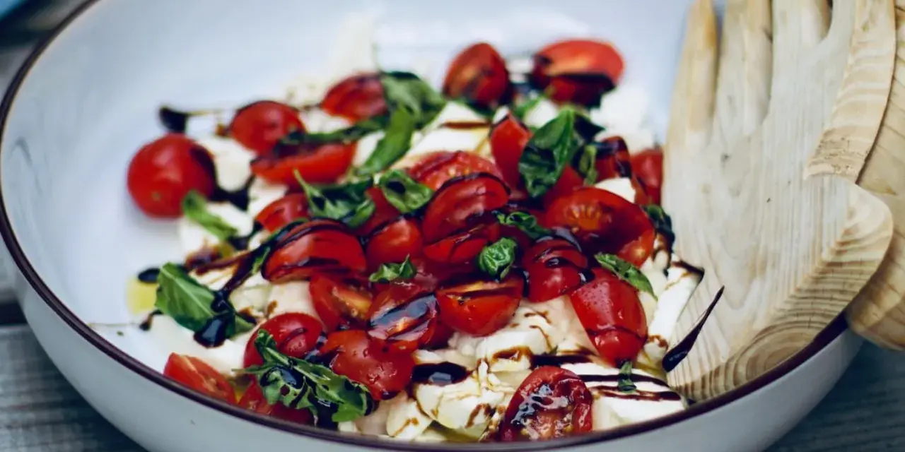 Burrata Caprese Salat mit Kirschtomaten, Basilikum und Balsamico-Dressing, serviert in einer Schüssel mit Holzsalatbesteck.