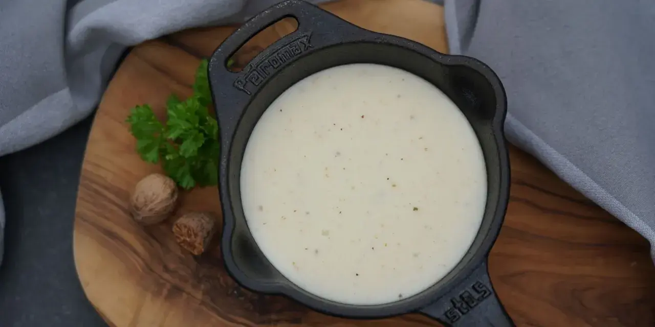 Cremige Bechamelsauce in einer Gusseisenpfanne, bereit für die Lasagne. Muskatnuss und Petersilie deuten auf die feinen Aromen hin.
