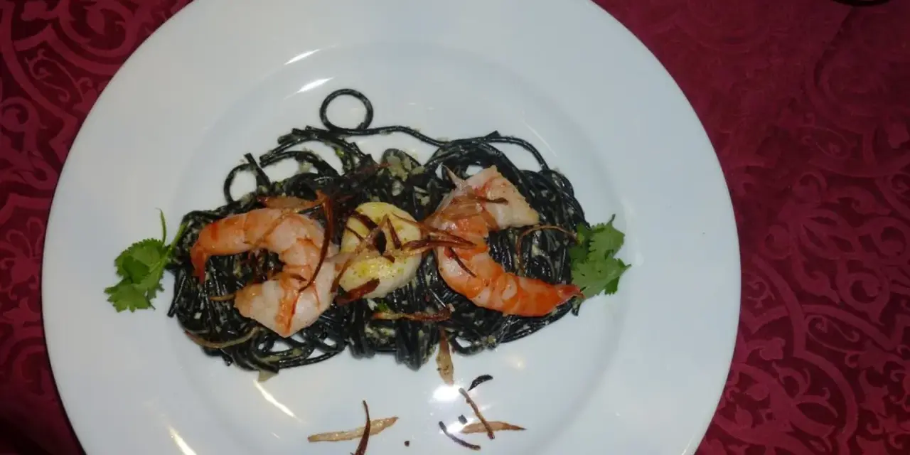 Schwarze Spaghetti mit Garnelen, garniert mit Koriander und knusprigen Zwiebeln. Ein köstliches Gericht, das auf einem weißen Teller serviert wird.