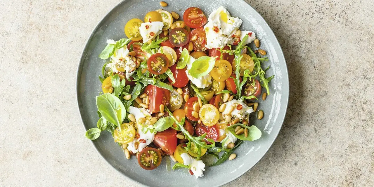 Bunter Tomatensalat mit cremiger Burrata, Rucola und Pinienkernen, garniert mit Basilikum.