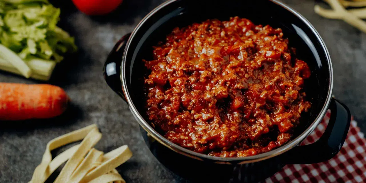 Herzhafte Bolognese-Sauce in einem Topf, daneben frische Bolognese-Zutaten wie Karotten, Sellerie und Tomaten.