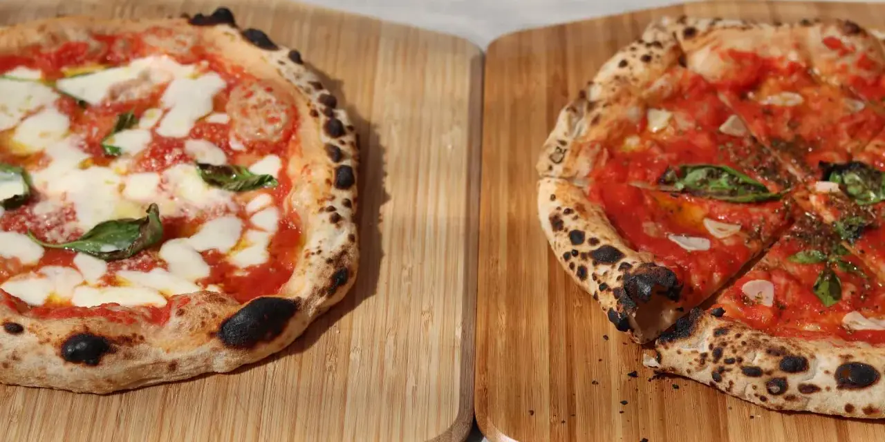 Zwei frisch gebackene Pizzen auf Holzbrettern, eine Margherita, die andere mit Tomaten und Basilikum.