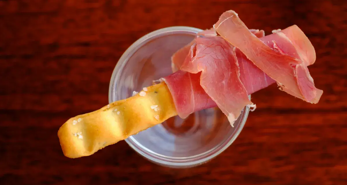 Ein knuspriger Grissini, umwickelt mit zartem Schinken, ruht in einem Glas. Perfekt für den kleinen Hunger.