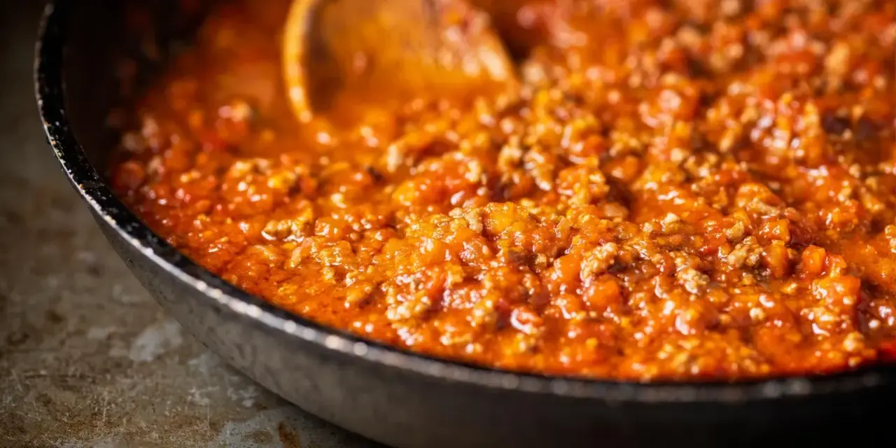 Ein Holzlöffel rührt in einer Pfanne mit köchelndem Sos bolognese.