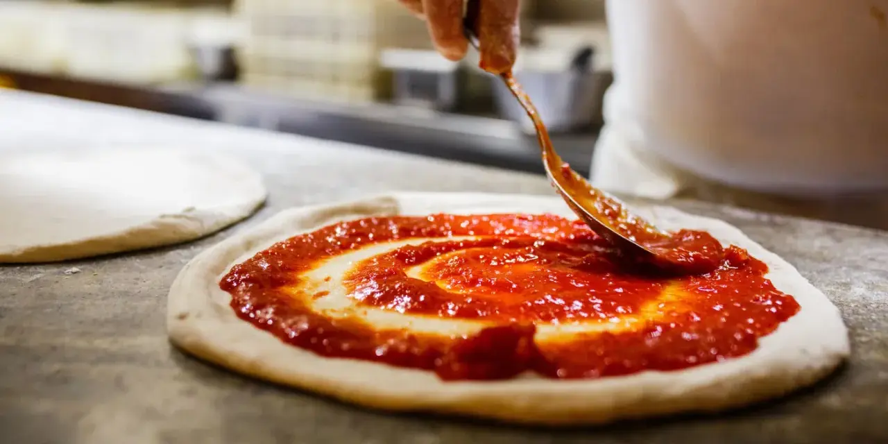 Eine Hand verteilt Tomatensauce auf einem Teigboden, die Vorbereitung für eine authentische pizza napoli original.