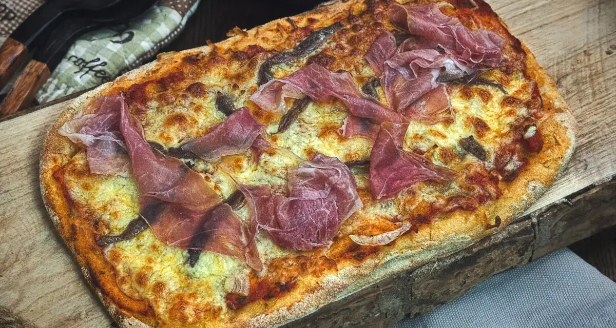 Eine rechteckige Pinsa mit geschmolzenem Käse und hauchdünnen Scheiben Schinken belegen. Köstlich!