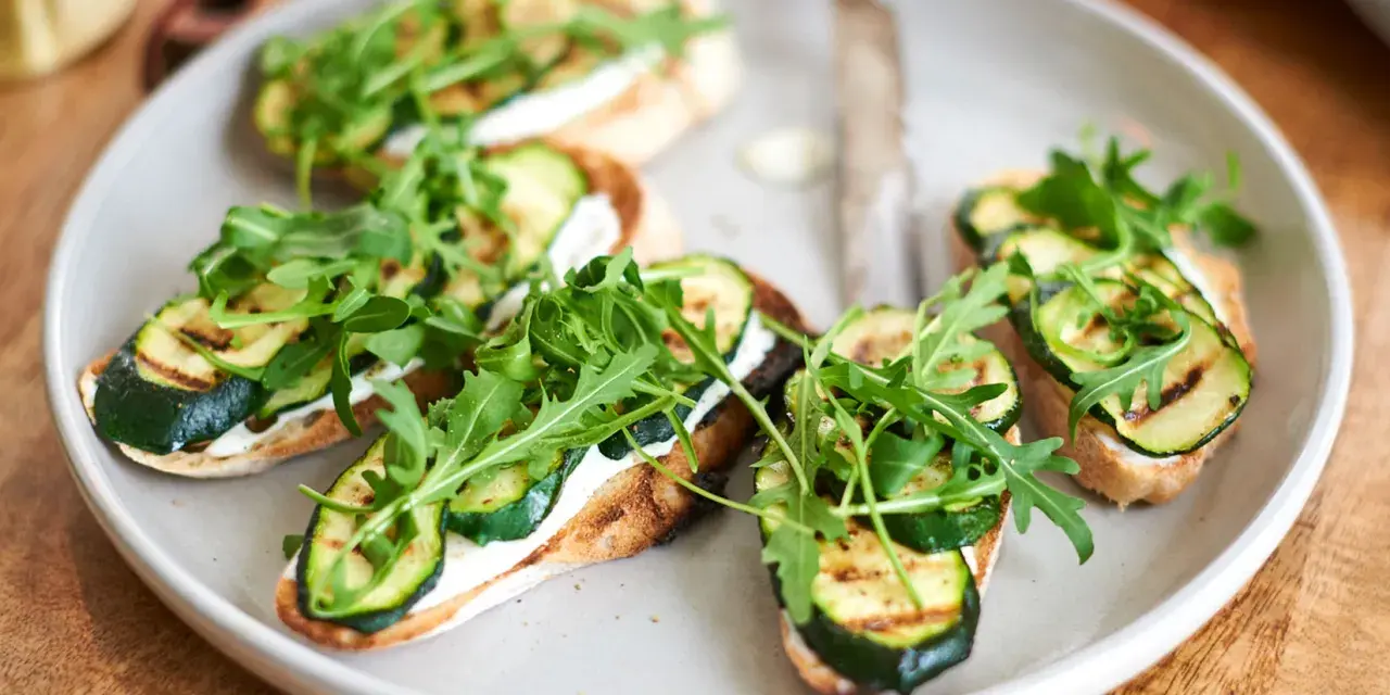 Italienische Crostini mit gegrillter Zucchini, Frischkäse und Rucola auf einem Teller.
