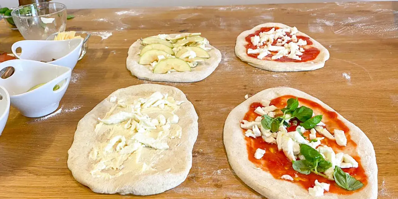 Vier vorbereitete Pinsa-Teiglinge liegen auf einem Holztisch. Einer mit Apfelscheiben, einer mit Tomatensoße und Mozzarella, einer nur mit Mozzarella und einer mit Tomatensoße, Mozzarella und Basilikum.