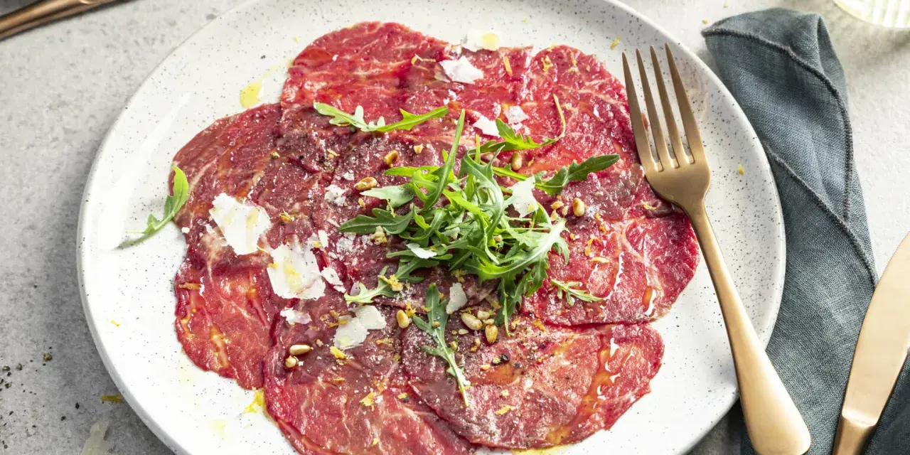 Carpaccio selber machen: hauchdünne Rinderfiletscheiben mit Rucola, Parmesan und Pinienkernen auf einem Teller.