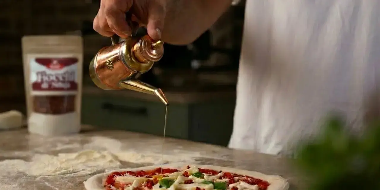 Ein Mann übergießt mit Olivenöl einen **Waldi Pizzateig**. Im Hintergrund eine Tüte mit Chiliflocken.