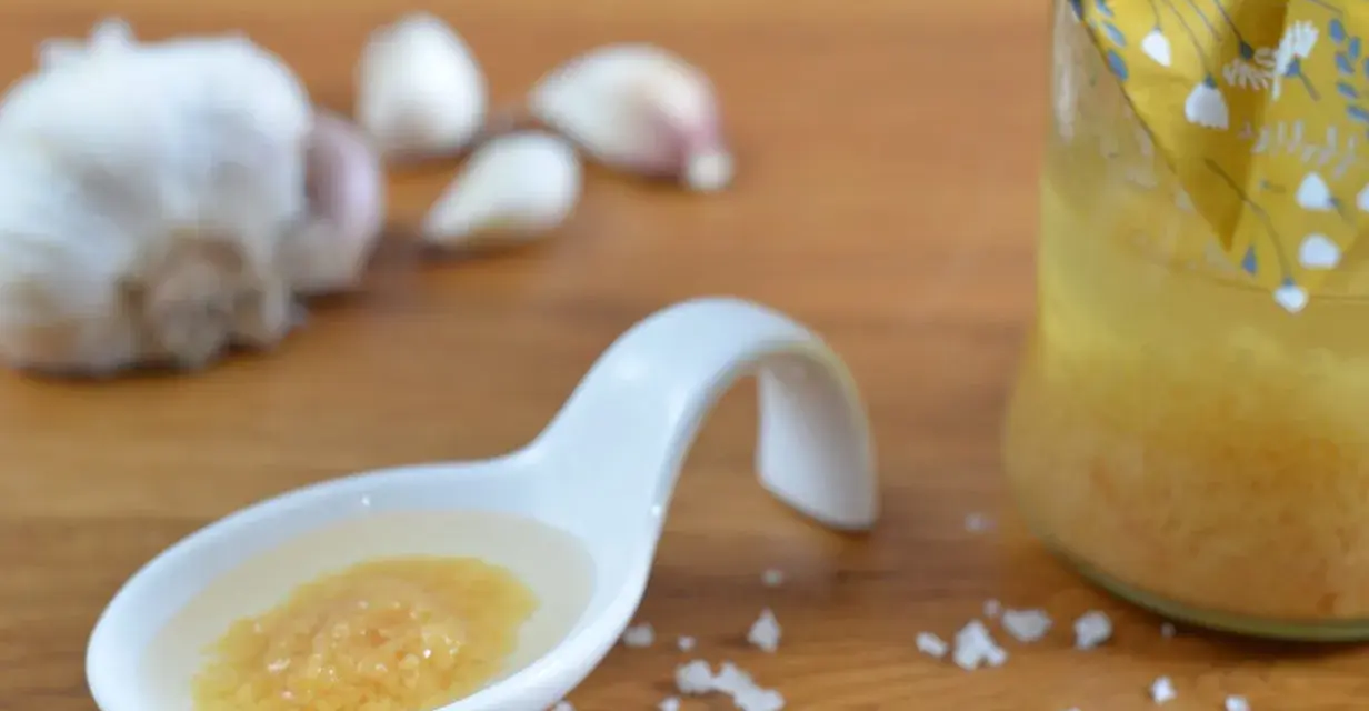 Ein Löffel mit einer köstlichen Knoblauchpaste, die an italienische Aromen erinnert. Daneben eine Flasche mit einer gelben Flüssigkeit und Knoblauchzehen.