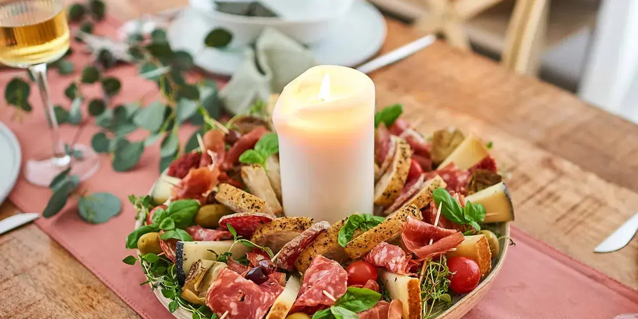 Ein festliches Arrangement mit italienischen Snacks zum Wein: Salami, Prosciutto, Käse, Oliven und Brot, umrahmt von Kräutern und einer Kerze.