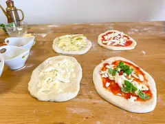 Vier vorbereitete Pinsa-Teiglinge liegen auf einem Holztisch. Einer mit Apfelscheiben, einer mit Tomatensoße und Mozzarella, einer nur mit Mozzarella und einer mit Tomatensoße, Mozzarella und Basilikum.