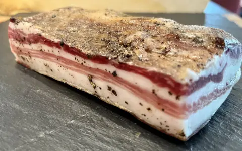 Ein Stück roher Pancetta mit deutlichen Schichten aus Fleisch und Fett, bestreut mit schwarzem Pfeffer. Perfekt, um ihn roh zu genießen.