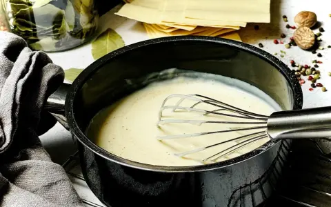 Cremige béchamelsauce mit Sahne wird in einem Topf mit einem Schneebesen zubereitet. Daneben liegen Lasagneplatten und Gewürze.