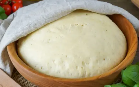 Ein aufgegangener Pizzateig in einer Holzschüssel, bedeckt mit einem Tuch. Die Frage "wie lange pizzateig gehen lassen" wird hier visuell beantwortet.