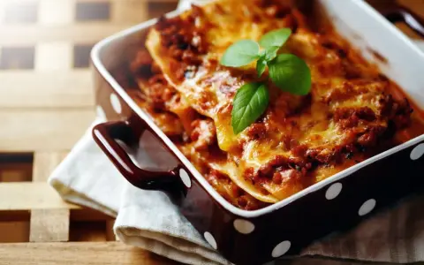 Herzhafte Lasagne Bolognese, eine der beliebtesten Lasagne Arten, dampft in einer braunen Auflaufform mit weißen Punkten. Frisch garniert mit Basilikum.
