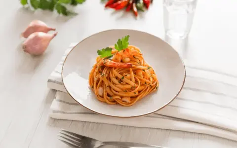 Ein Teller mit Linguini Nudeln mit Garnelen und Tomatensauce, garniert mit Petersilie. Daneben Schalotten, Chilis und ein Glas Wasser.
