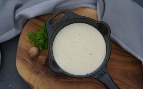 Cremige Bechamelsauce in einer Gusseisenpfanne, bereit für die Lasagne. Muskatnuss und Petersilie deuten auf die feinen Aromen hin.