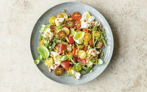 Bunter Tomatensalat mit cremiger Burrata, Rucola und Pinienkernen, garniert mit Basilikum.