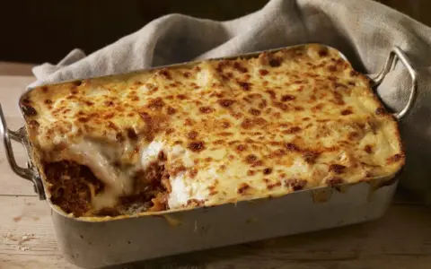 Herzhafte Lasagne mit goldbraun überbackener Käseschicht und einem Hauch von Creme Fraiche, serviert in einer Metallform auf Holztisch.