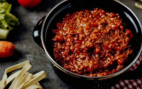 Herzhafte Bolognese-Sauce in einem Topf, daneben frische Bolognese-Zutaten wie Karotten, Sellerie und Tomaten.