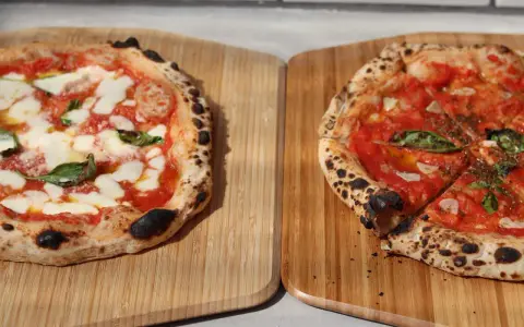 Zwei frisch gebackene Pizzen auf Holzbrettern, eine Margherita, die andere mit Tomaten und Basilikum.