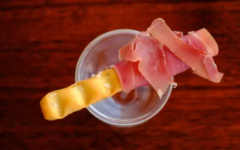 Ein knuspriger Grissini, umwickelt mit zartem Schinken, ruht in einem Glas. Perfekt für den kleinen Hunger.
