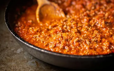 Ein Holzlöffel rührt in einer Pfanne mit köchelndem Sos bolognese.