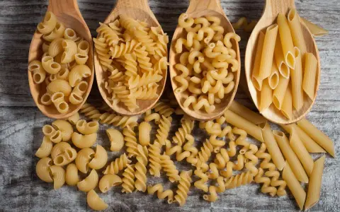 Vier Holzlöffel mit verschiedenen Nudelsorten: Maccheroni, Fusilli, Cavatappi und Penne. Die perfekte Menge Nudeln pro Person für ein köstliches Essen.
