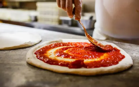 Eine Hand verteilt Tomatensauce auf einem Teigboden, die Vorbereitung für eine authentische pizza napoli original.