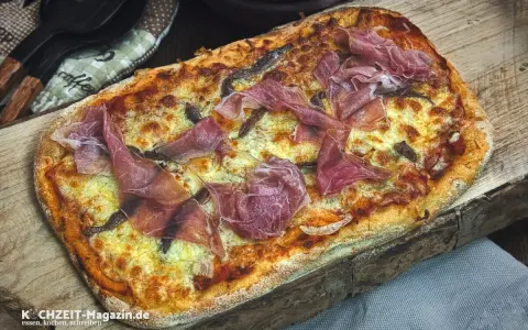 Eine rechteckige Pinsa mit geschmolzenem Käse und hauchdünnen Scheiben Schinken belegen. Köstlich!